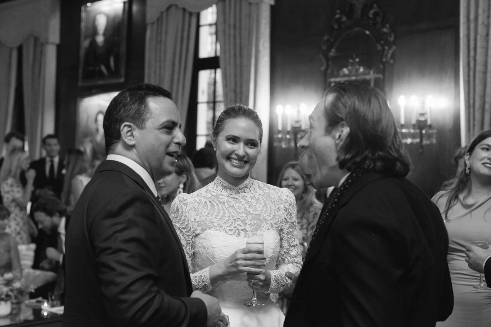 People at a formal event, three individuals engaged in conversation, including a woman in a white lace dress holding a glass, in an elegant room with wood-paneled walls and candlelit lighting.