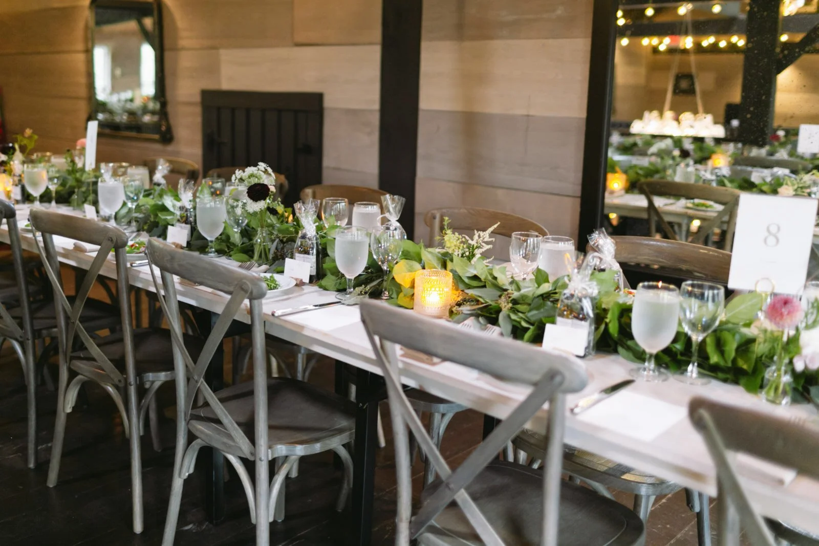 A long wedding reception table decorated with a lush green foliage runner, flowers, candles, and place settings with plates, silverware, and glasses in a rustic wooden room with warm lighting.