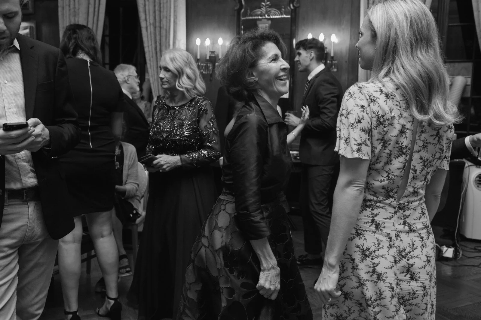 People socializing at an indoor event or party, with women talking and smiling, some in formal attire, in a decorated room with warm lighting.