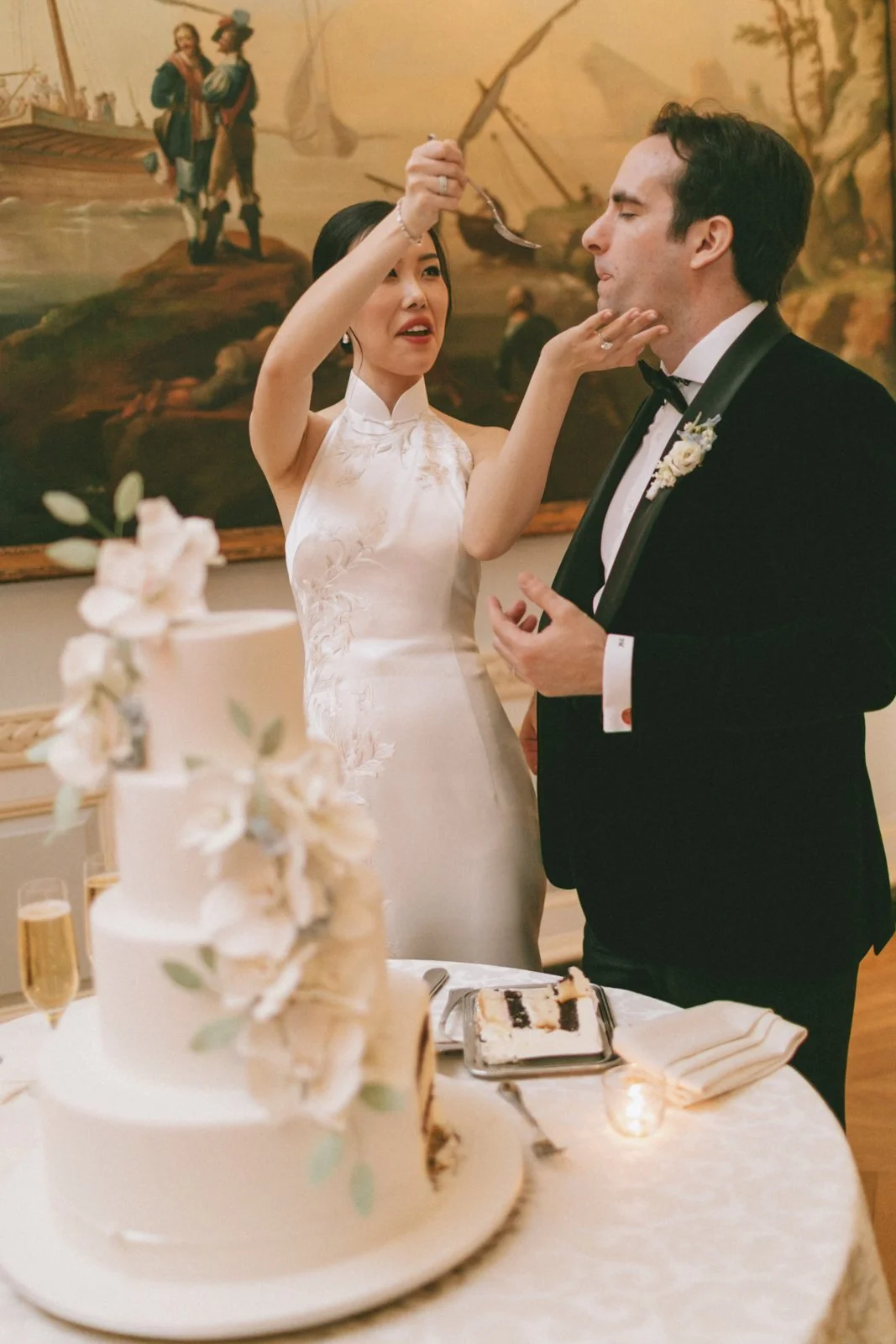 A bride and groom during a wedding reception, with the bride feeding the groom a piece of cake, surrounded by a layered wedding cake with flowers on top, and a painting in the background.