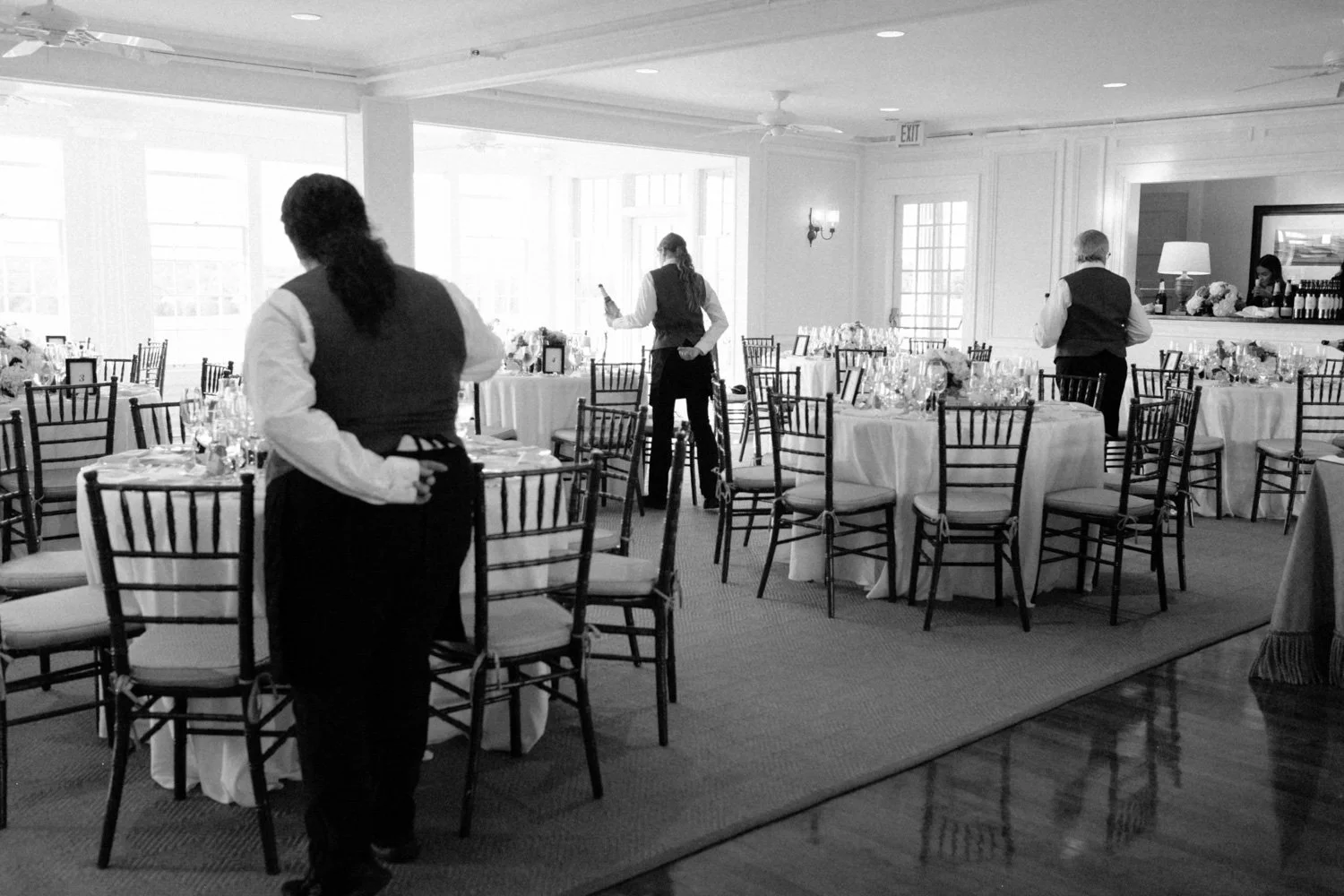 A banquet hall with round tables covered in white tablecloths, set with glassware and floral centerpieces, and black chairs. Three servers are preparing the room, one in the foreground with a hand behind their back, and two others in the background a
