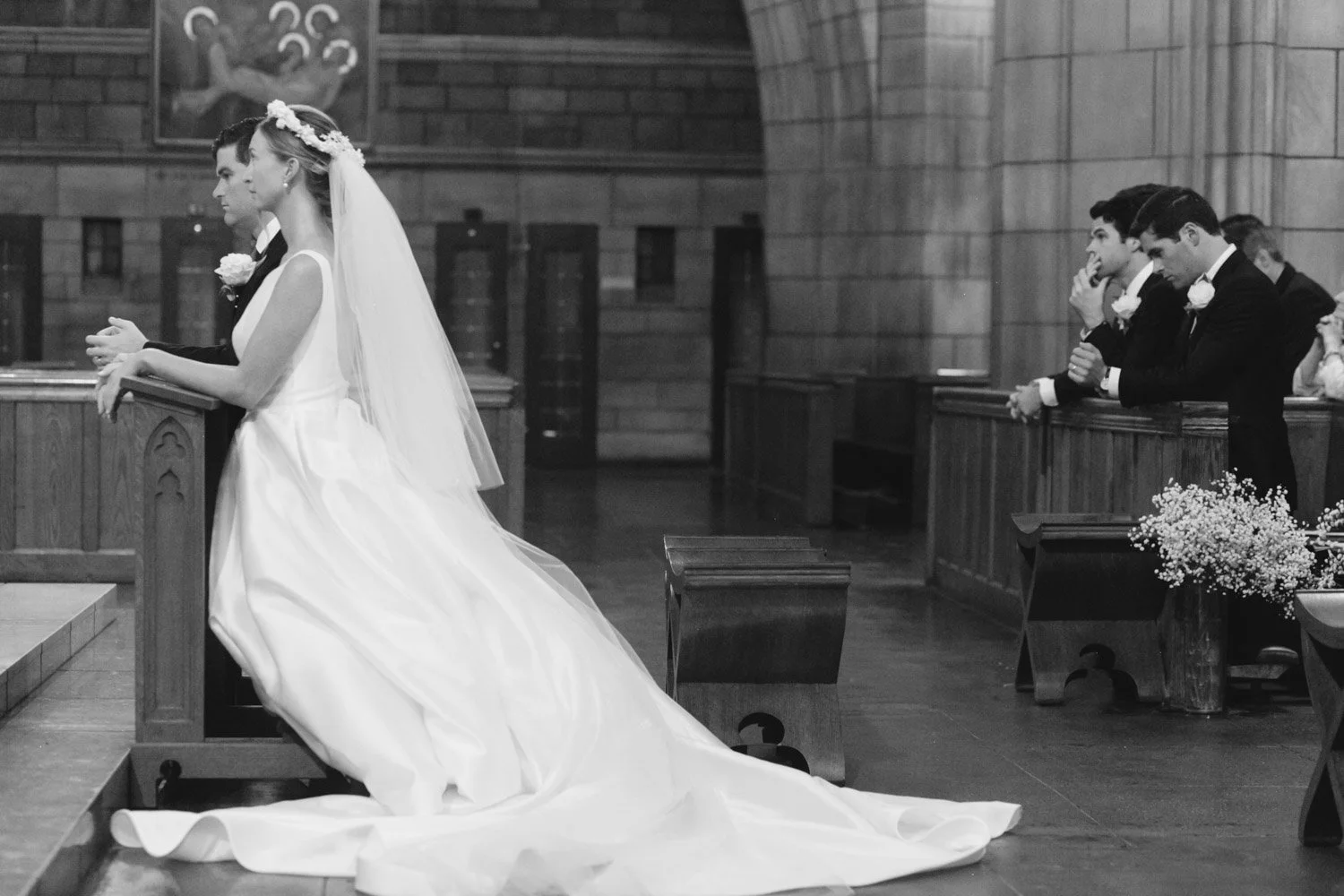 A bride and groom kneel in prayer at the altar during a wedding ceremony inside a church, with bridesmaids and groomsmen praying nearby.