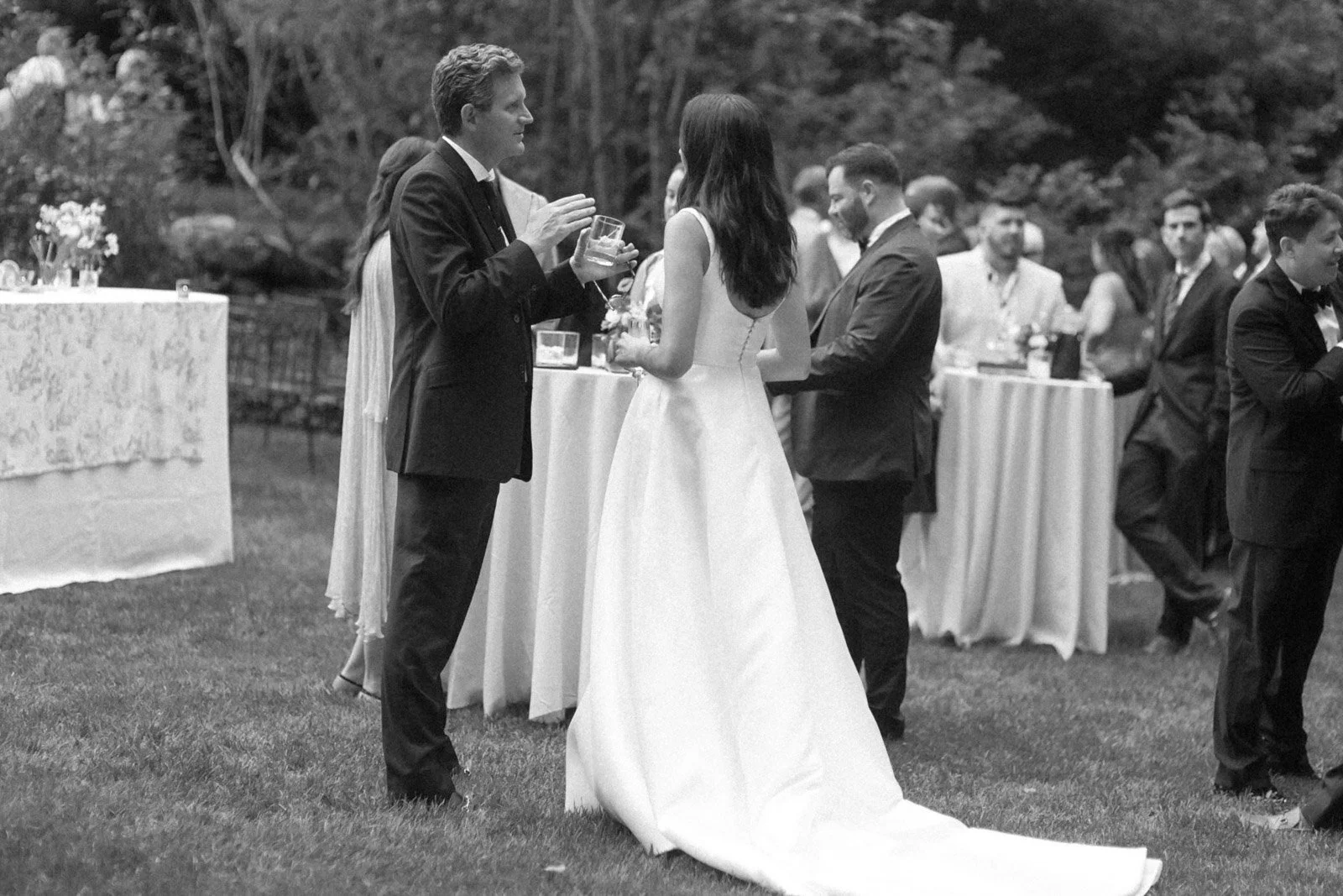Black and white photo of a wedding reception outdoors showing a bride in a white gown talking to a groom in a black tuxedo. There are other guests in formal attire in the background gathered around high tables.