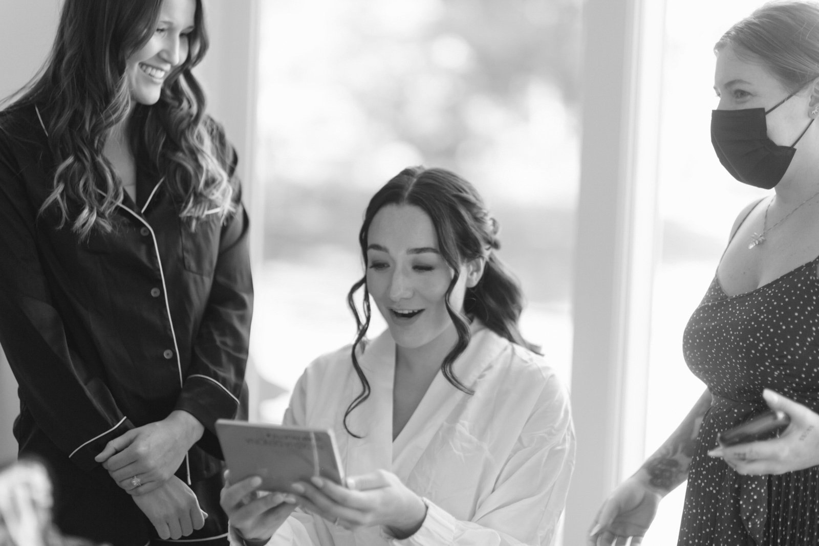 A woman in a white robe looks surprised and happy while looking at her phone, surrounded by two women, one in pajamas and the other in a polka-dot dress wearing a face mask, inside a well-lit room.