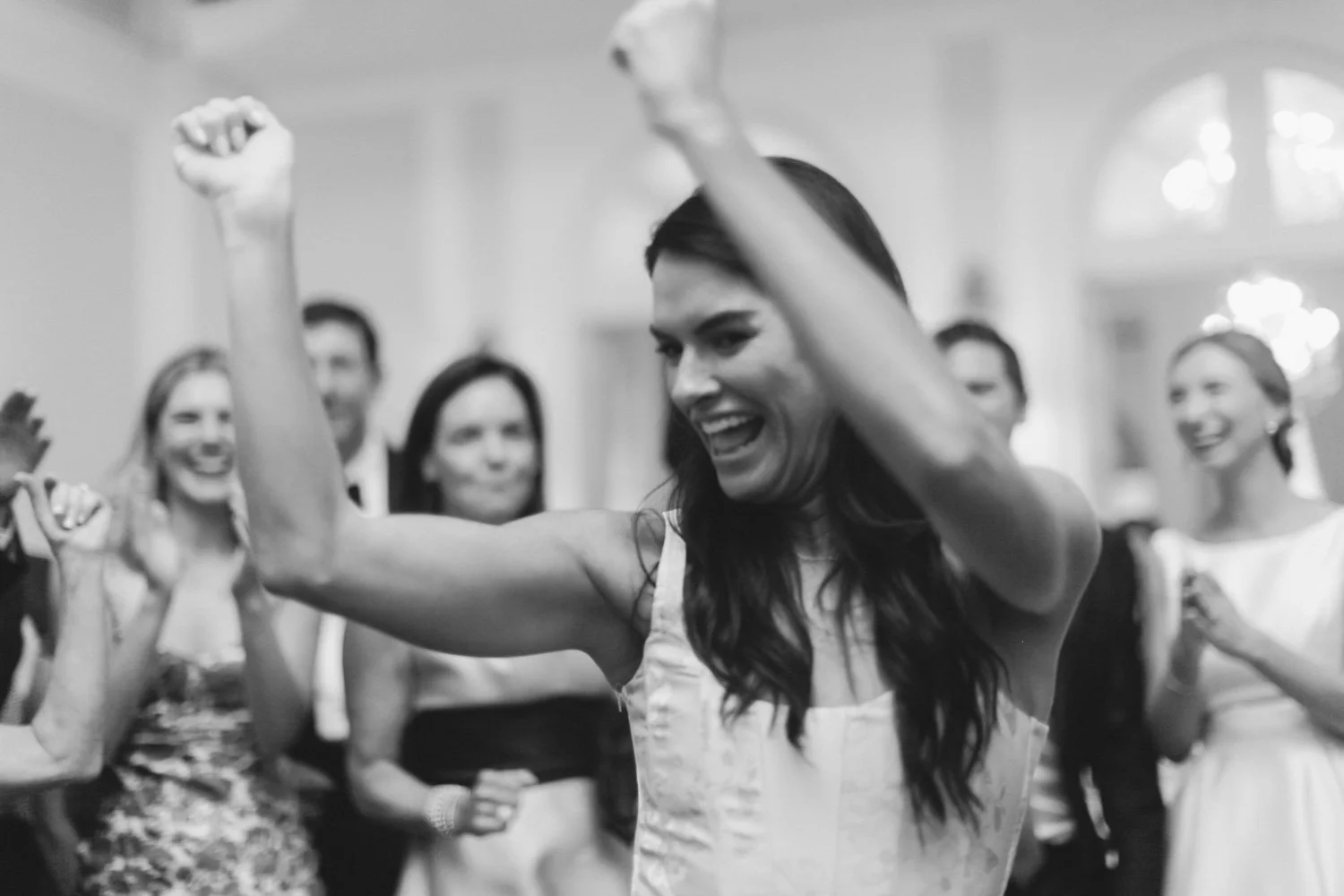 A woman in a sleeveless dress lifting her arms and smiling, surrounded by a group of women at a celebration in an elegant room.