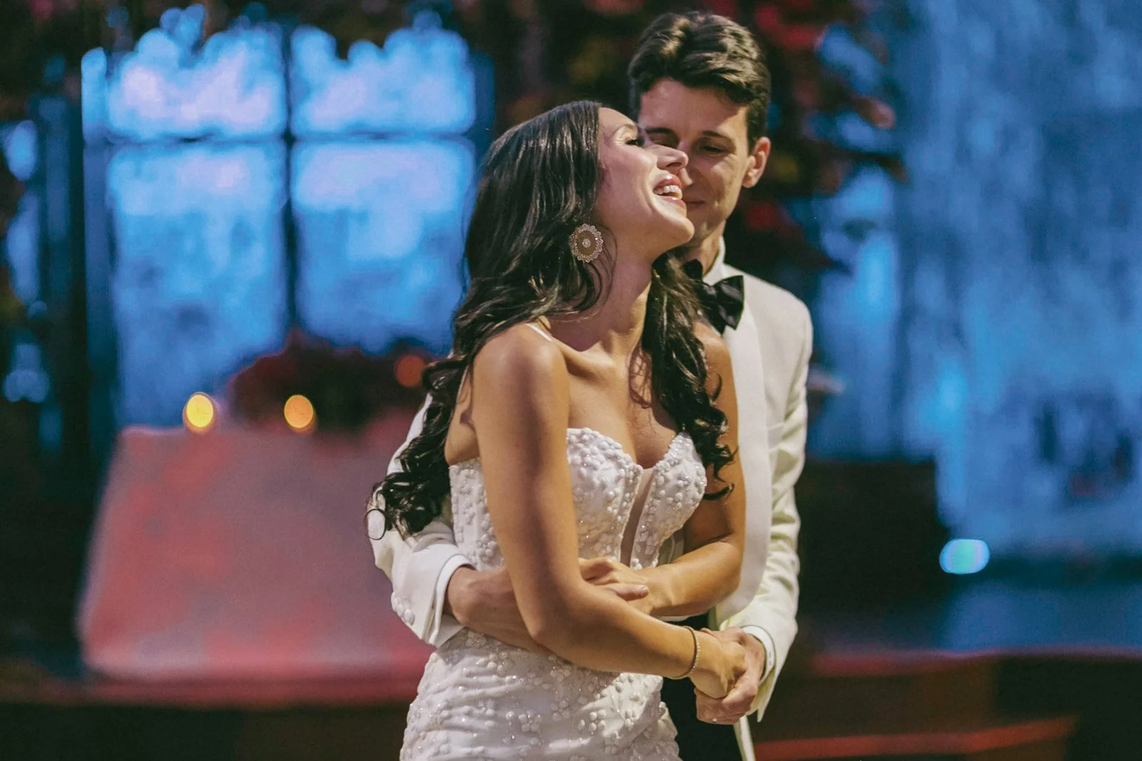 A woman in a white wedding dress and a man in a tuxedo are dancing closely, smiling and enjoying a romantic moment at a wedding reception.