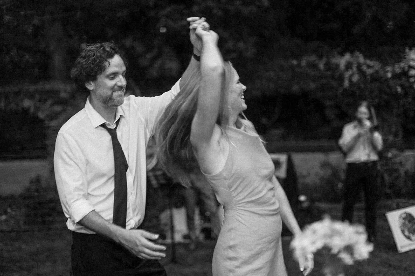 A man in a white shirt and black tie with a woman in a dress dancing outdoors at night, while another person stands in the background taking a photo.