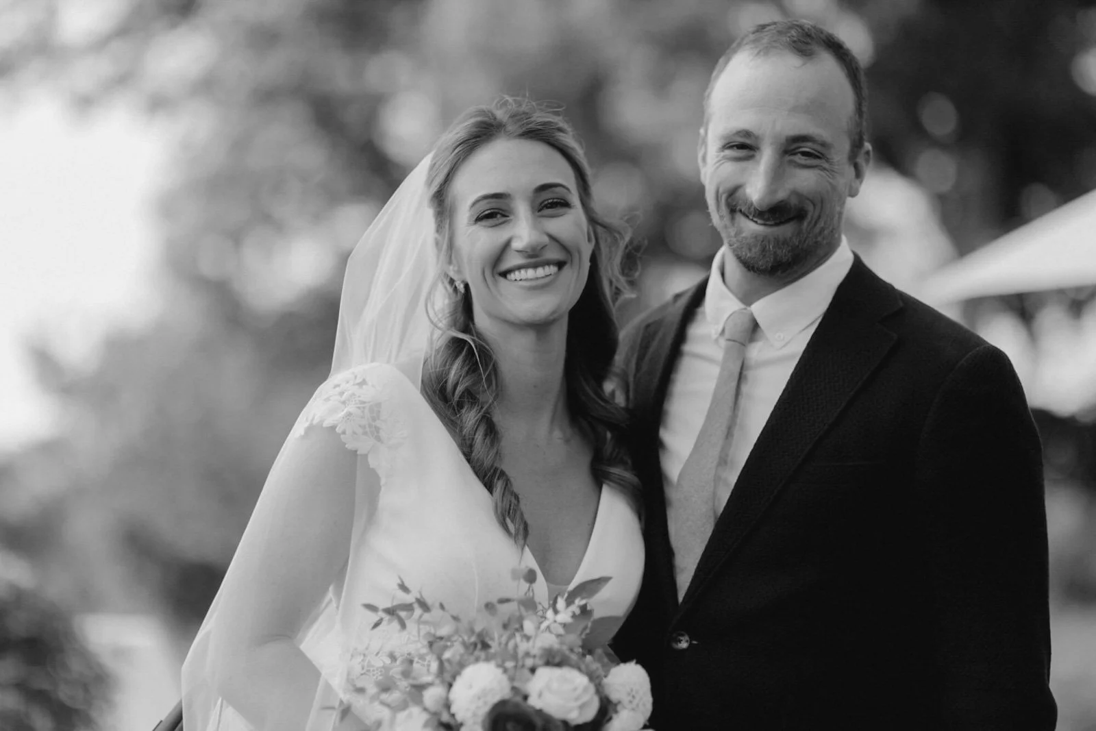 A smiling bride in a wedding dress and veil holding a bouquet stands next to a smiling groom in a suit outside.