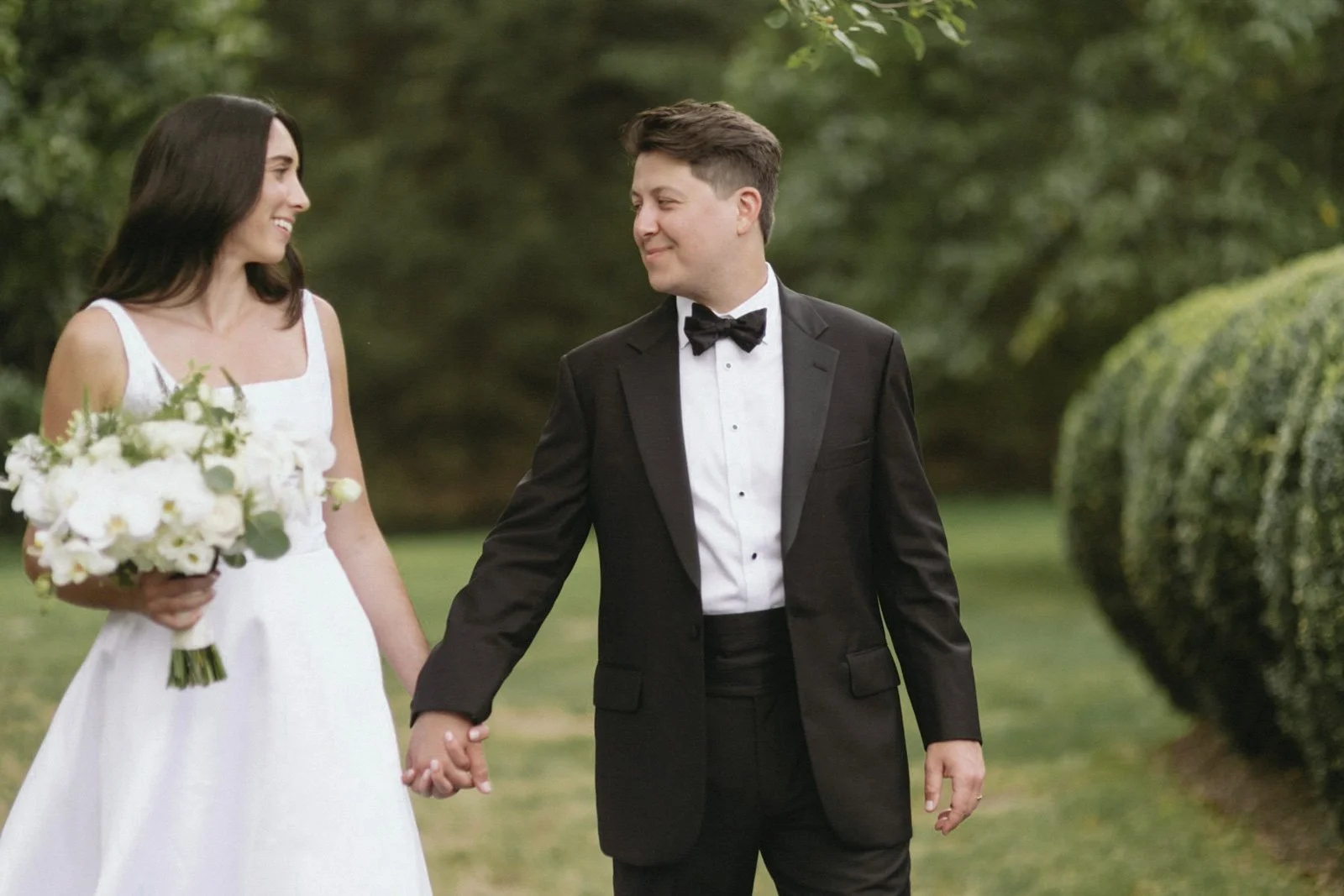 A bride and groom holding hands in an outdoor setting, with the bride wearing a white wedding dress and holding a bouquet of white flowers, and the groom wearing a black tuxedo with a bow tie, smiling at each other.