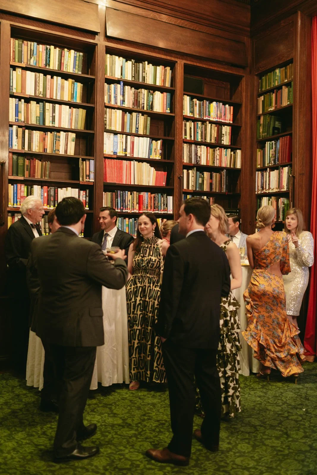 People at a social gathering in a library with floor-to-ceiling bookshelves filled with books, dressed in formal attire.