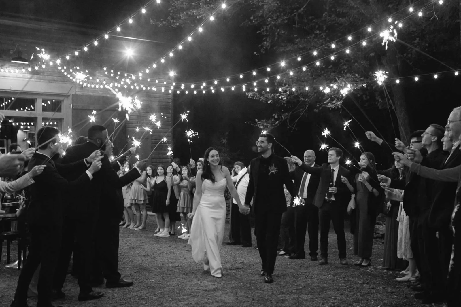 A black and white photo of a wedding celebration, with a bride and groom holding hands walking through a crowd of guests holding sparklers and string lights overhead.