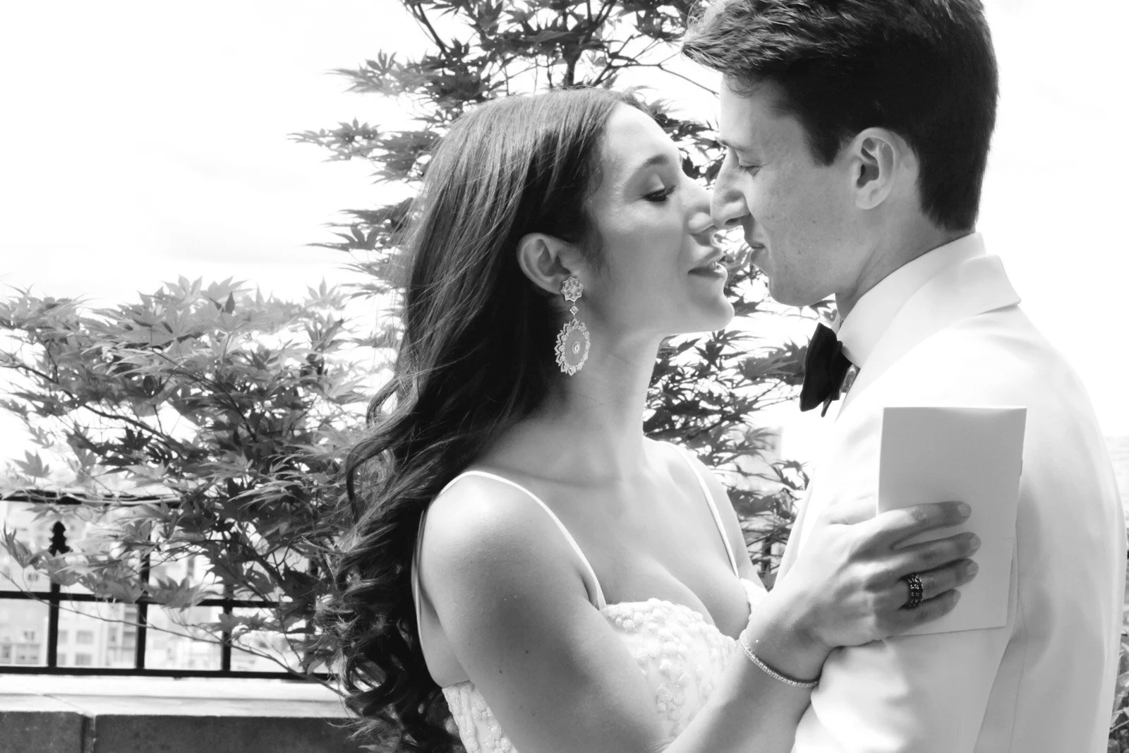 A bride and groom in wedding attire sharing a close moment outdoors, with trees and buildings in the background.