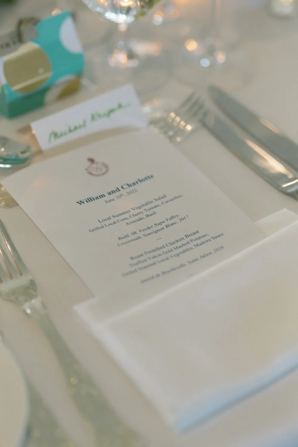 A wedding dinner table setting with a printed menu, silverware, a folded white napkin, and a name tag reading "Amanda Jane." The menu features dishes like a vegetable salad and chicken breast, with decorative lights in the background.
