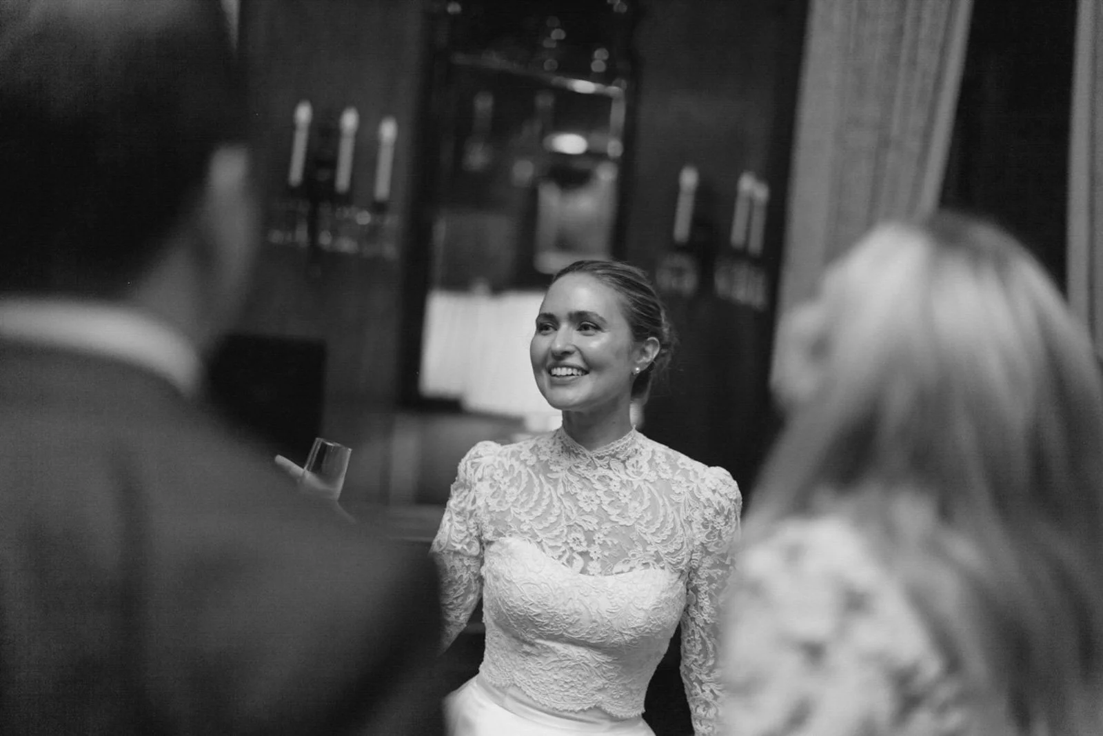 A smiling woman in a lace dress at a formal event, seen from the front, with blurred people in the foreground.