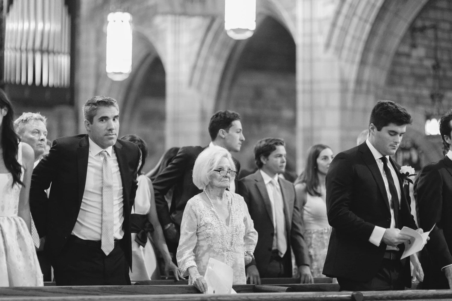 People standing in church pews during a formal ceremony inside a church with high vaulted ceilings and hanging lights.
