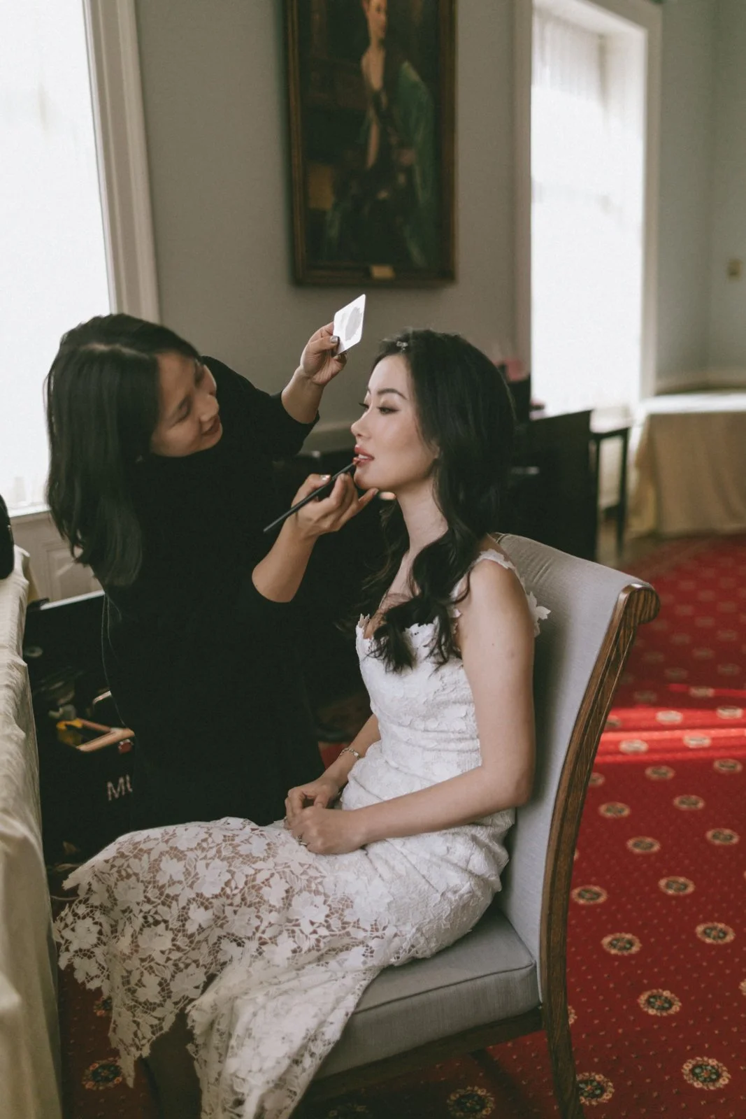 A woman in a white lace dress is getting her makeup done by a makeup artist in an elegant room, with a portrait on the wall and a red carpeted floor.