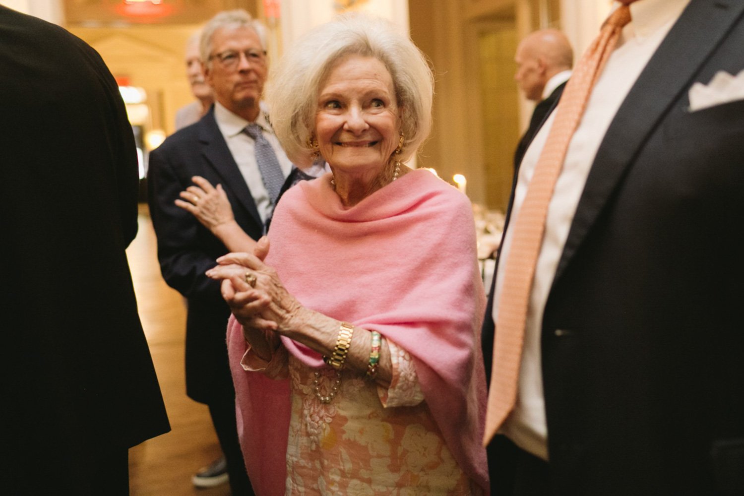 An elderly woman smiling and holding hands with a man at a social gathering, wearing a pink shawl and jewelry, surrounded by other older adults in formal attire.