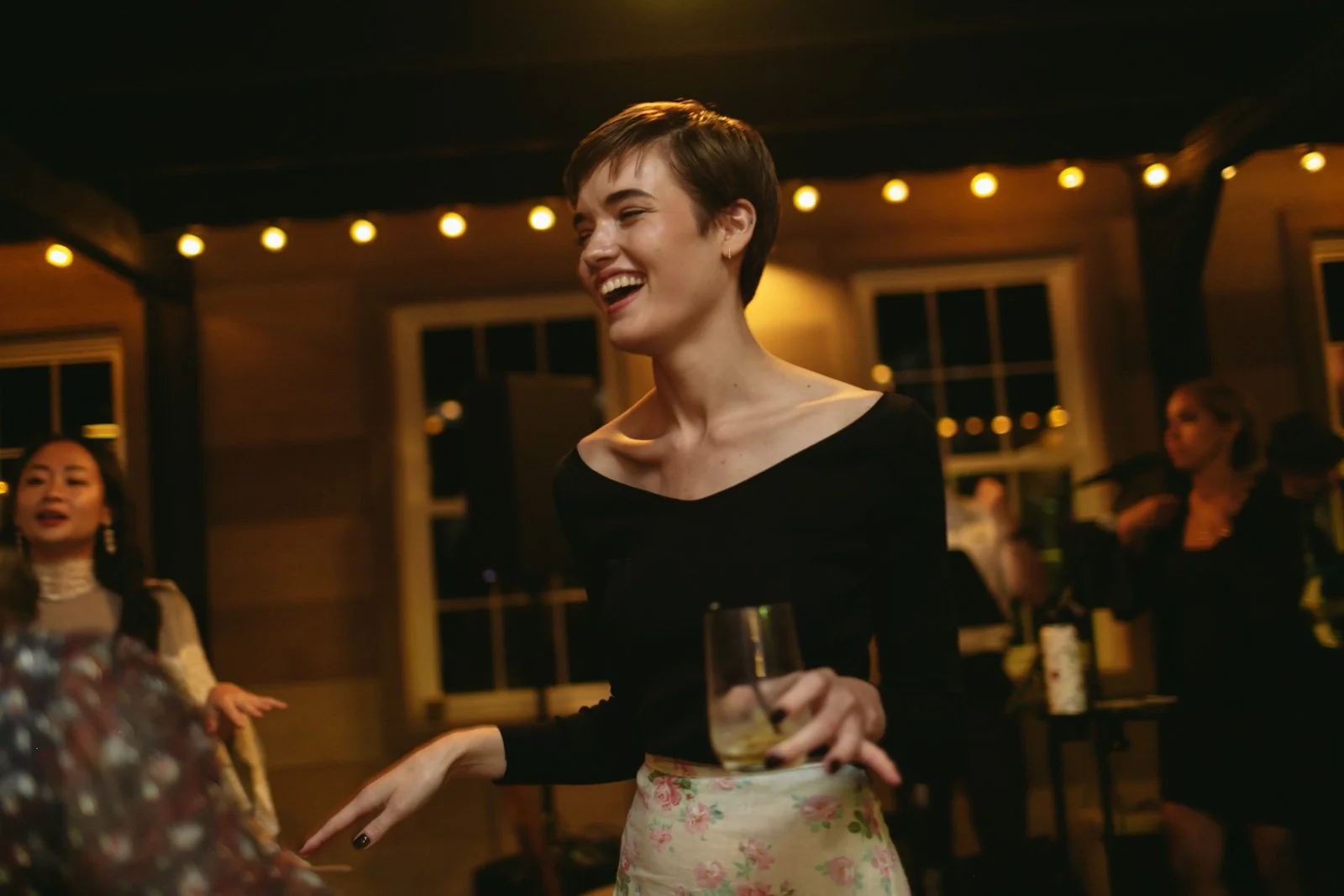 A woman with short brown hair wearing a black off-the-shoulder top and a floral skirt, smiling and holding a drink at a party or social gathering in a warmly lit wooden venue.