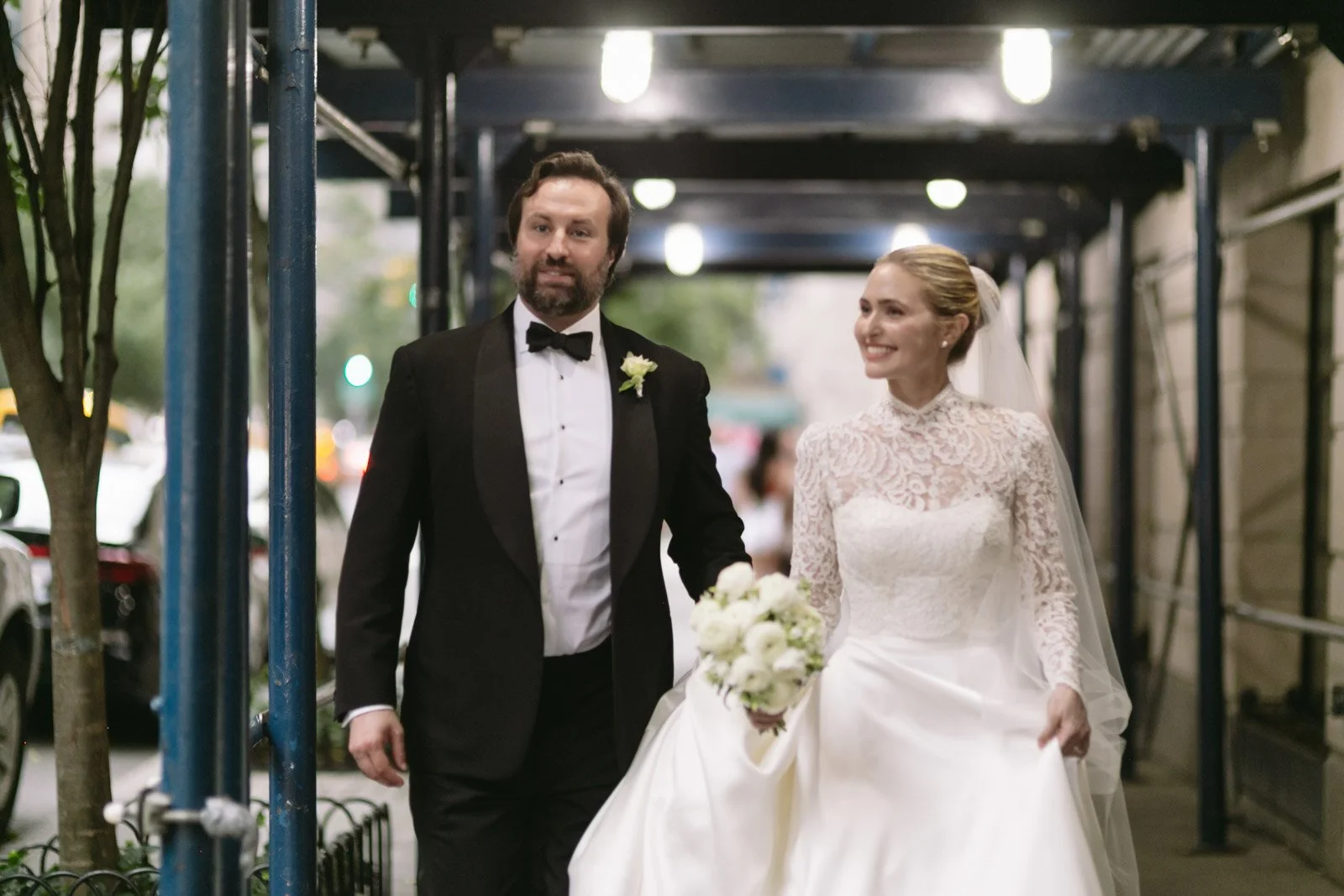 Bride and groom walking on city sidewalk during wedding, the groom in a tuxedo and the bride in a wedding dress holding a bouquet.
