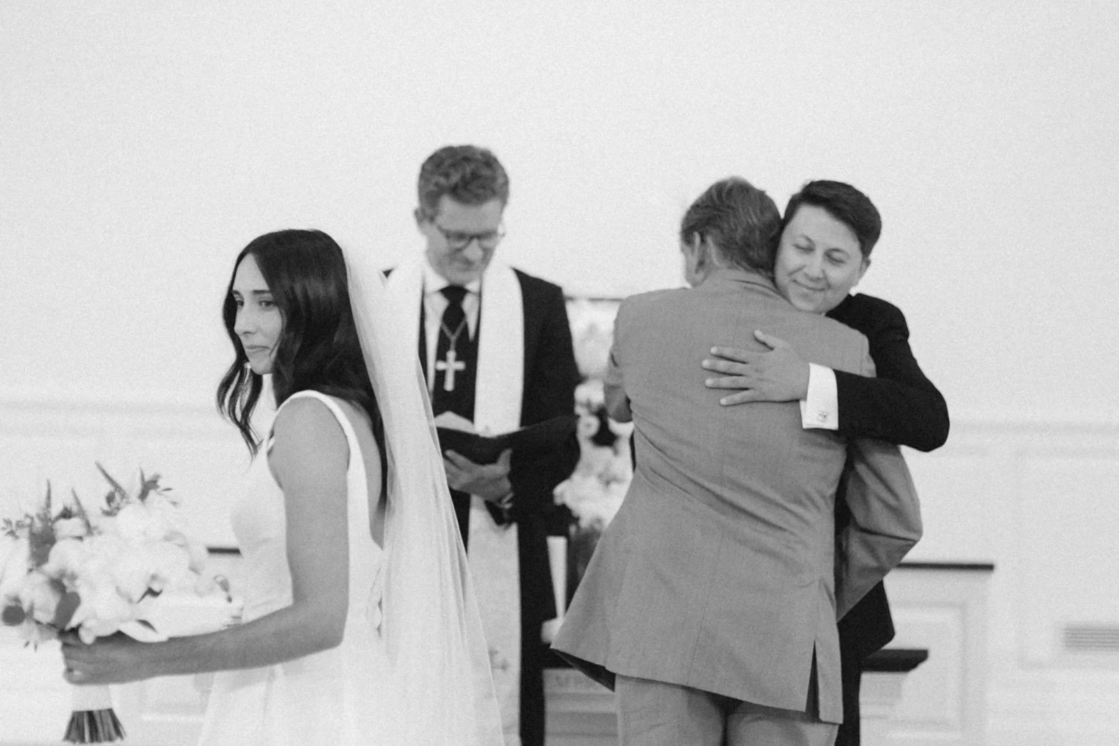 A black-and-white photo of a wedding ceremony with a bride holding a bouquet on the left and two men hugging in an embrace on the right, while a pastor officiates in the background.