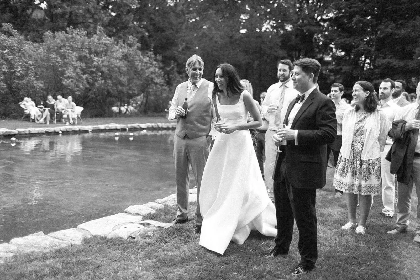 A group of elegantly dressed people at a wedding reception outdoors by a pond, with some guests seated on the far side, in a garden setting with trees.