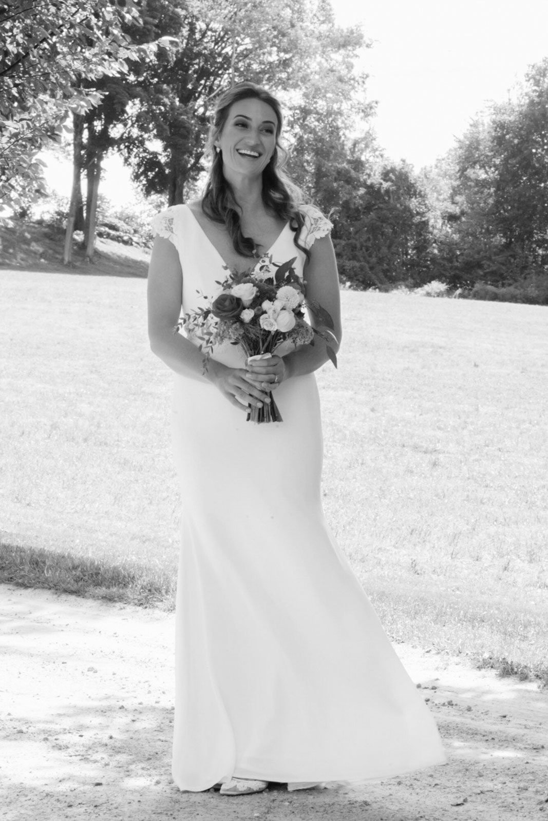 Black and white photo of a smiling woman in a wedding dress holding a bouquet, standing outdoors on a grassy area with trees in the background.