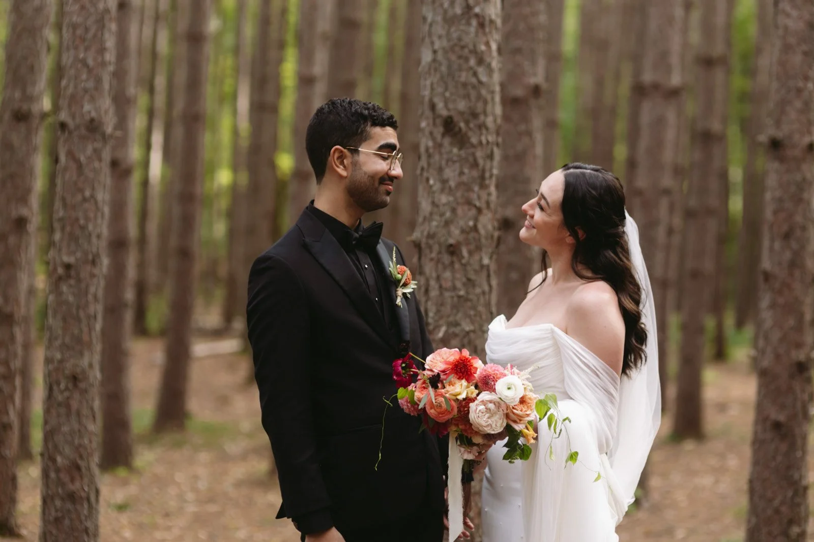 A bride and groom stand facing each other in a forest, smiling. The groom wears a black suit and glasses, holding a bouquet of pink and white flowers. The bride wears a white off-the-shoulder wedding dress with dark hair, holding a similar bouquet.