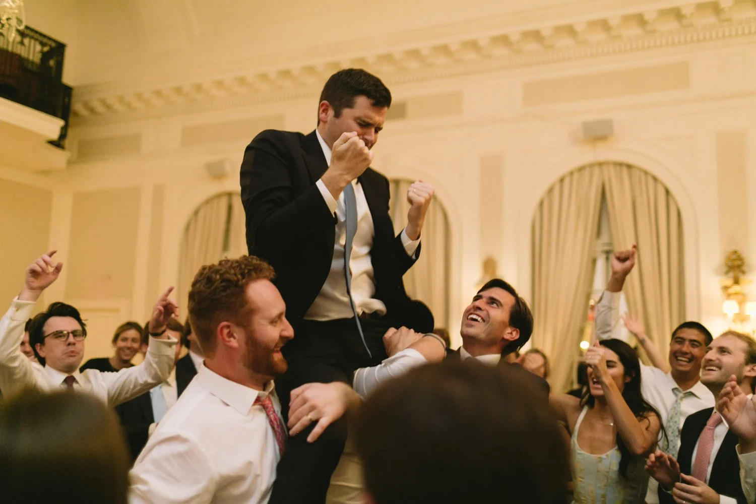 Man in a suit raising his fist in celebration while sitting on someone's shoulders at a lively party or celebration with smiling people around.