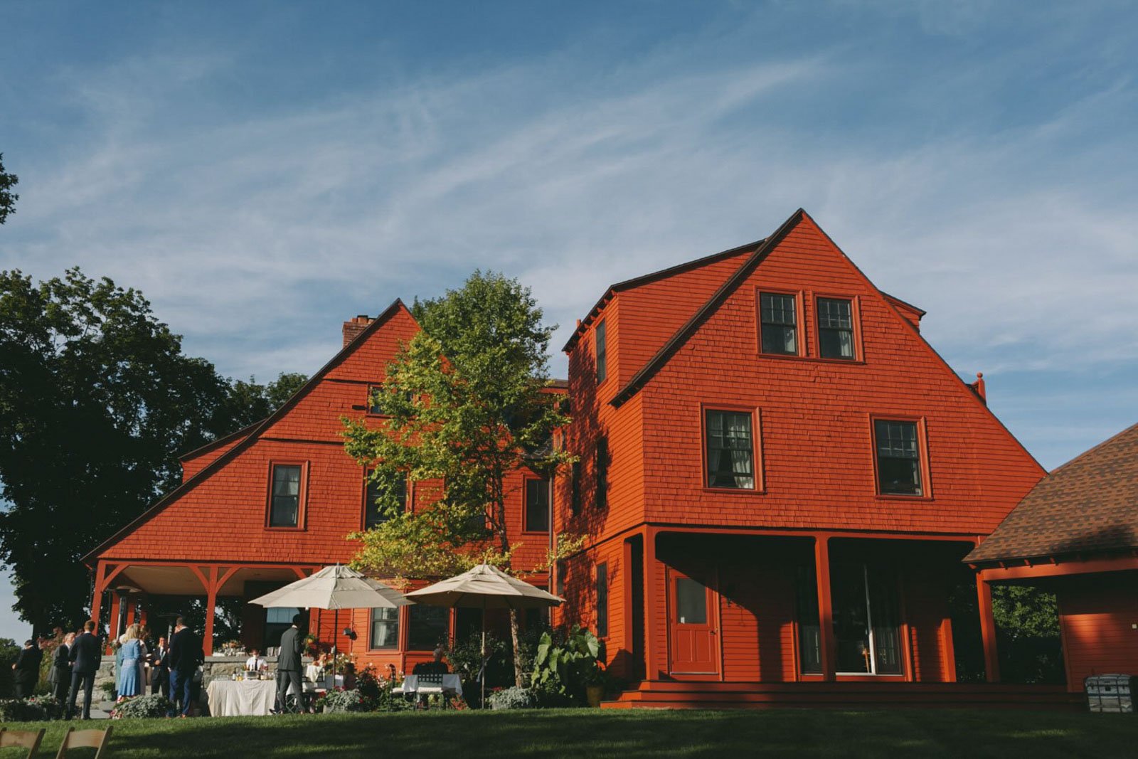 A large red wooden house with multiple stories, surrounded by trees, with a lawn and outdoor seating with umbrellas, and people gathered, possibly for an event.