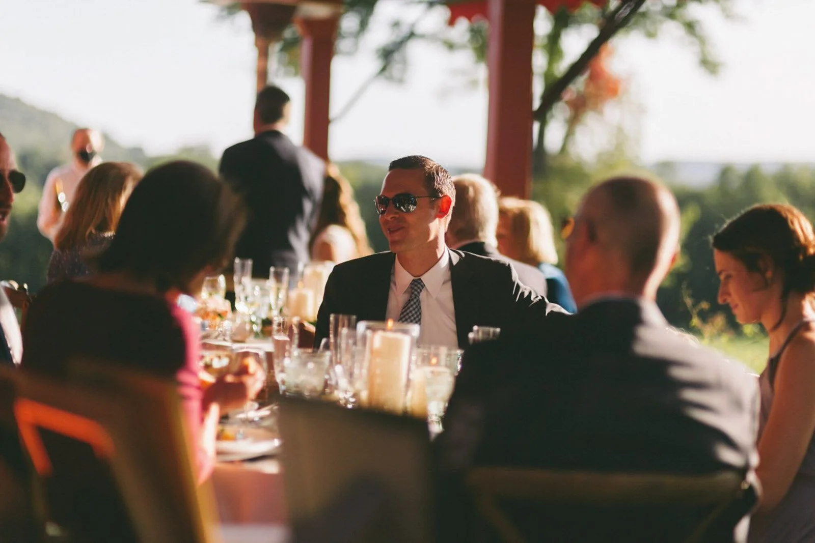 People sitting at a banquet table outdoors during daytime, dressed in formal attire, with a man in sunglasses at the center, smiling and engaged in conversation.