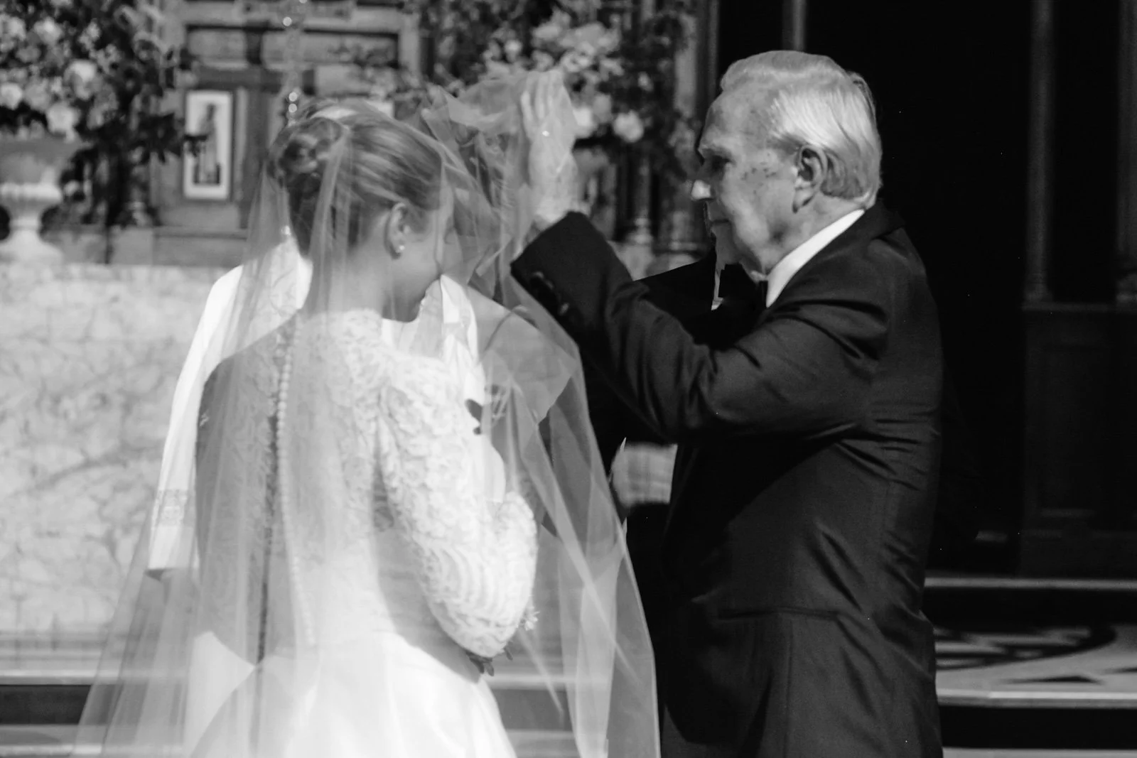 An elderly man in a tuxedo placing a veil on a young woman in a wedding dress during a wedding ceremony.
