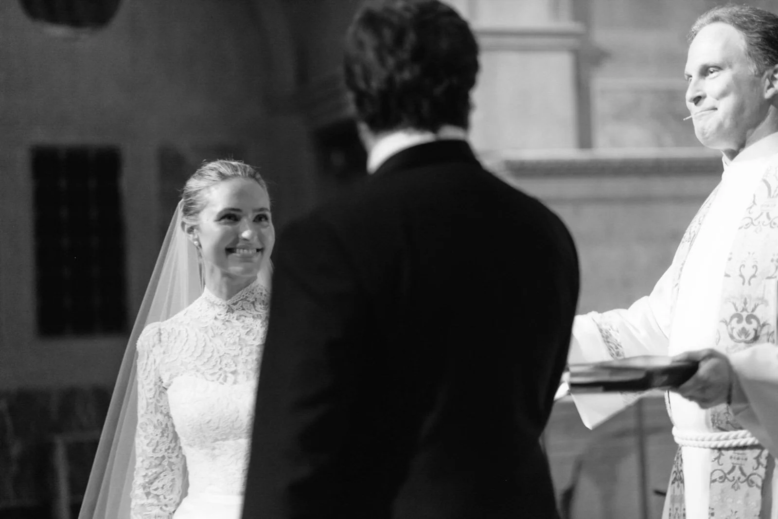 A wedding ceremony with a bride smiling at the groom, who is facing a priest in a church. The priest is holding a book or folder, and the bride is wearing a white lace wedding dress with a veil.