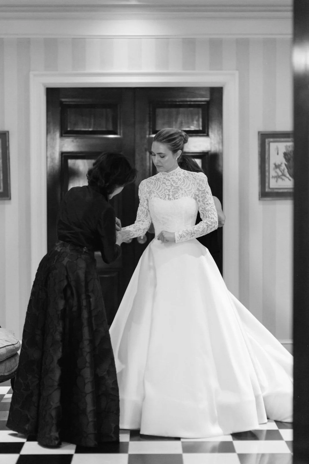 A woman in a wedding dress being assisted by another woman in a black dress, standing in a room with dark wood doors and framed pictures on the wall.