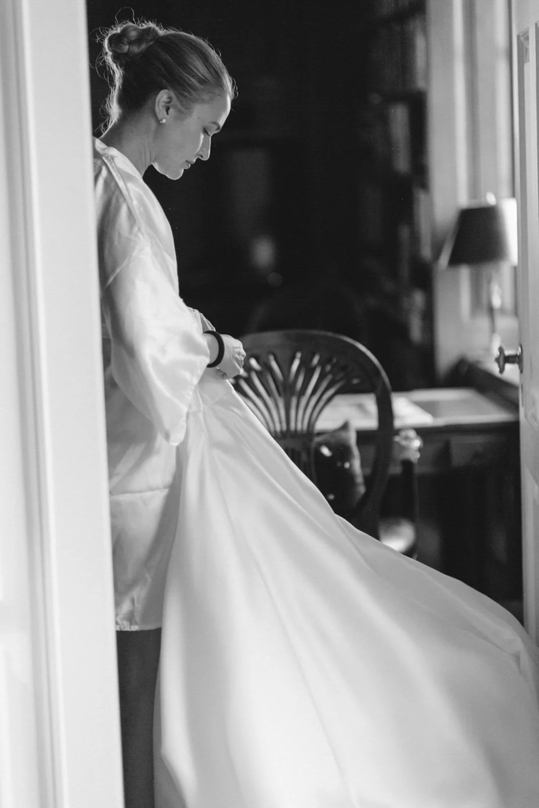 A woman in a silk robe adjusting her wedding dress in a room, viewed through a door frame, black and white photo.