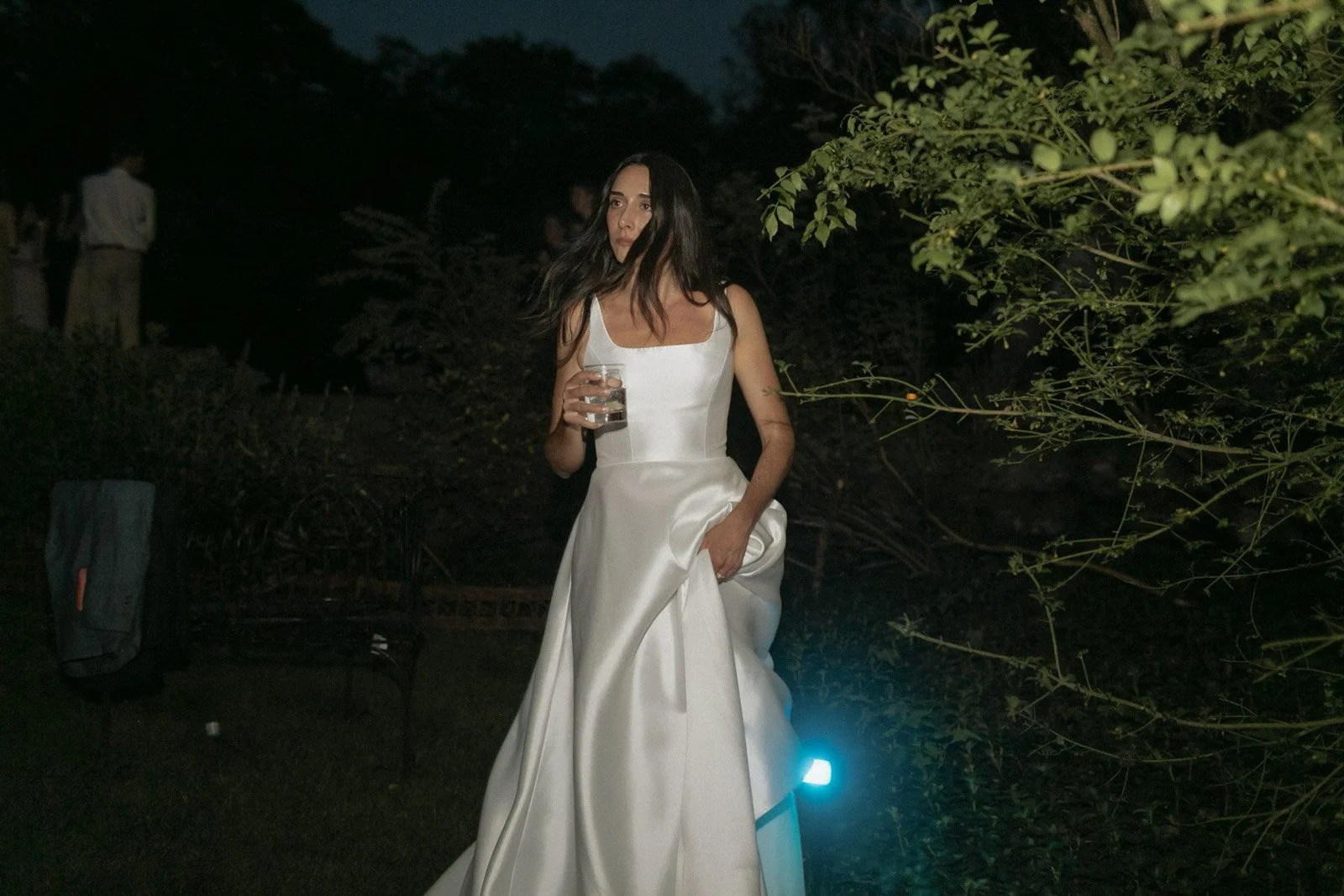 Woman in a white satin gown holding a glass of water at an outdoor evening event, surrounded by greenery and other figures in the background.