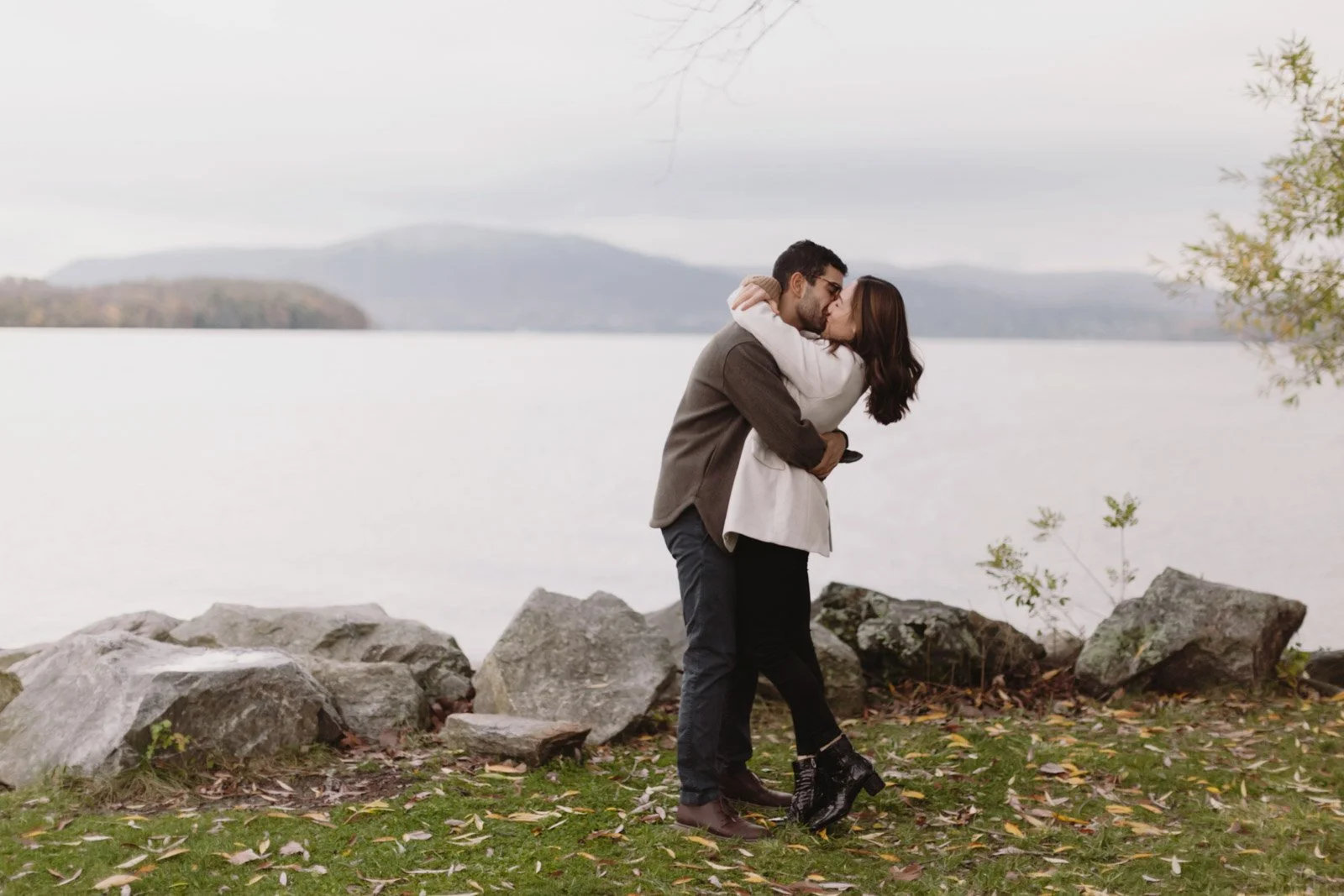 A couple embracing and kissing by a lakeside on a cloudy day, with rocks on the shore and distant hills in the background.