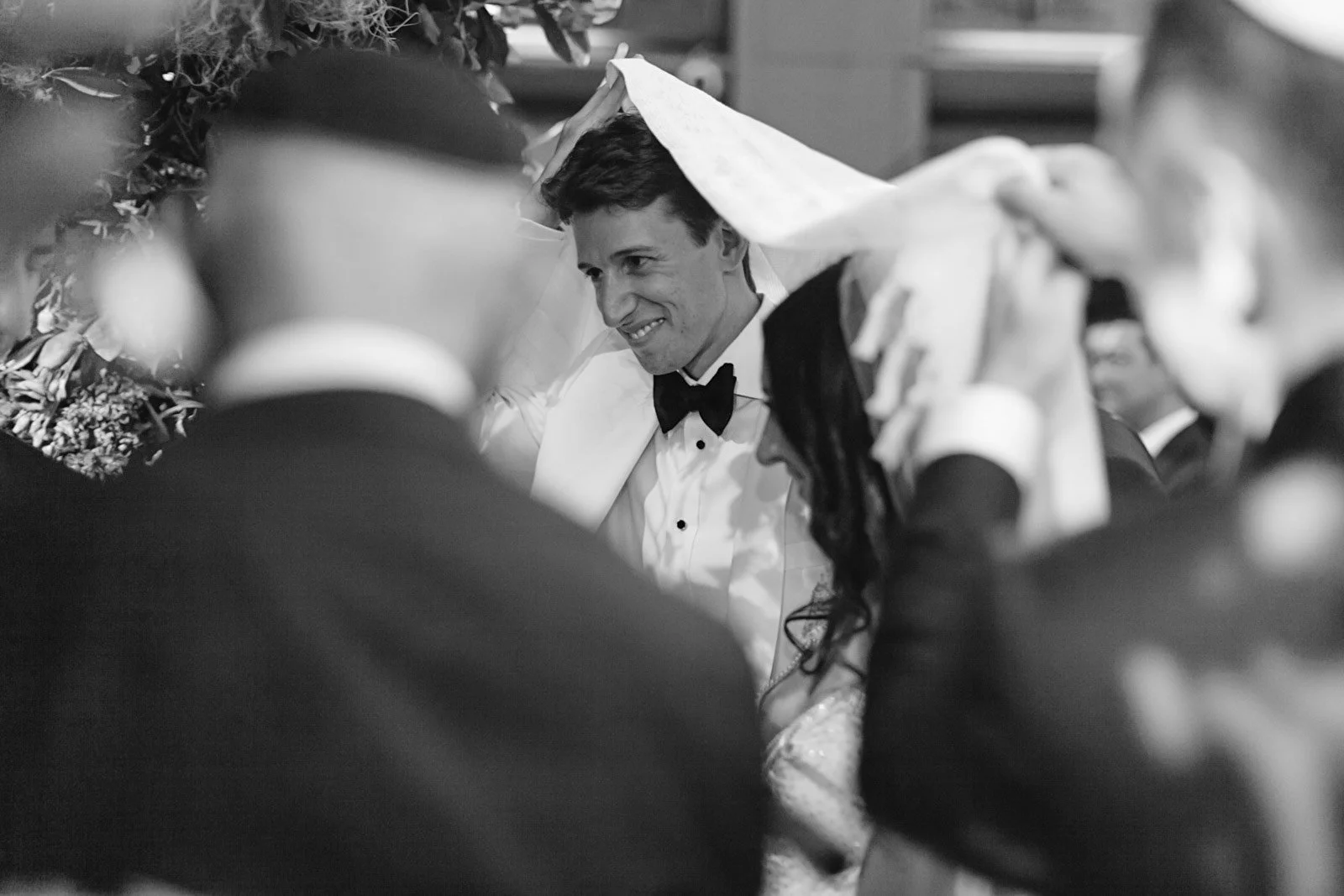 A black-and-white photo of a joyful man in a tuxedo with a bowtie, surrounded by people, seemingly during a wedding ceremony or celebration.