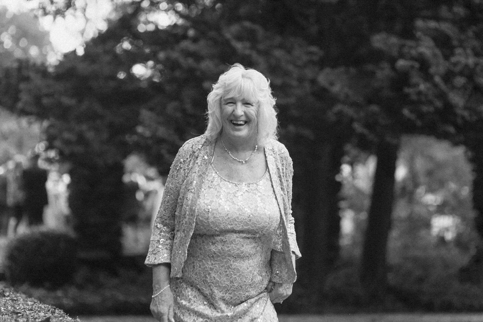 An elderly woman with curly hair, smiling and wearing a lace dress and jewelry, standing outdoors in a park with trees in the background.