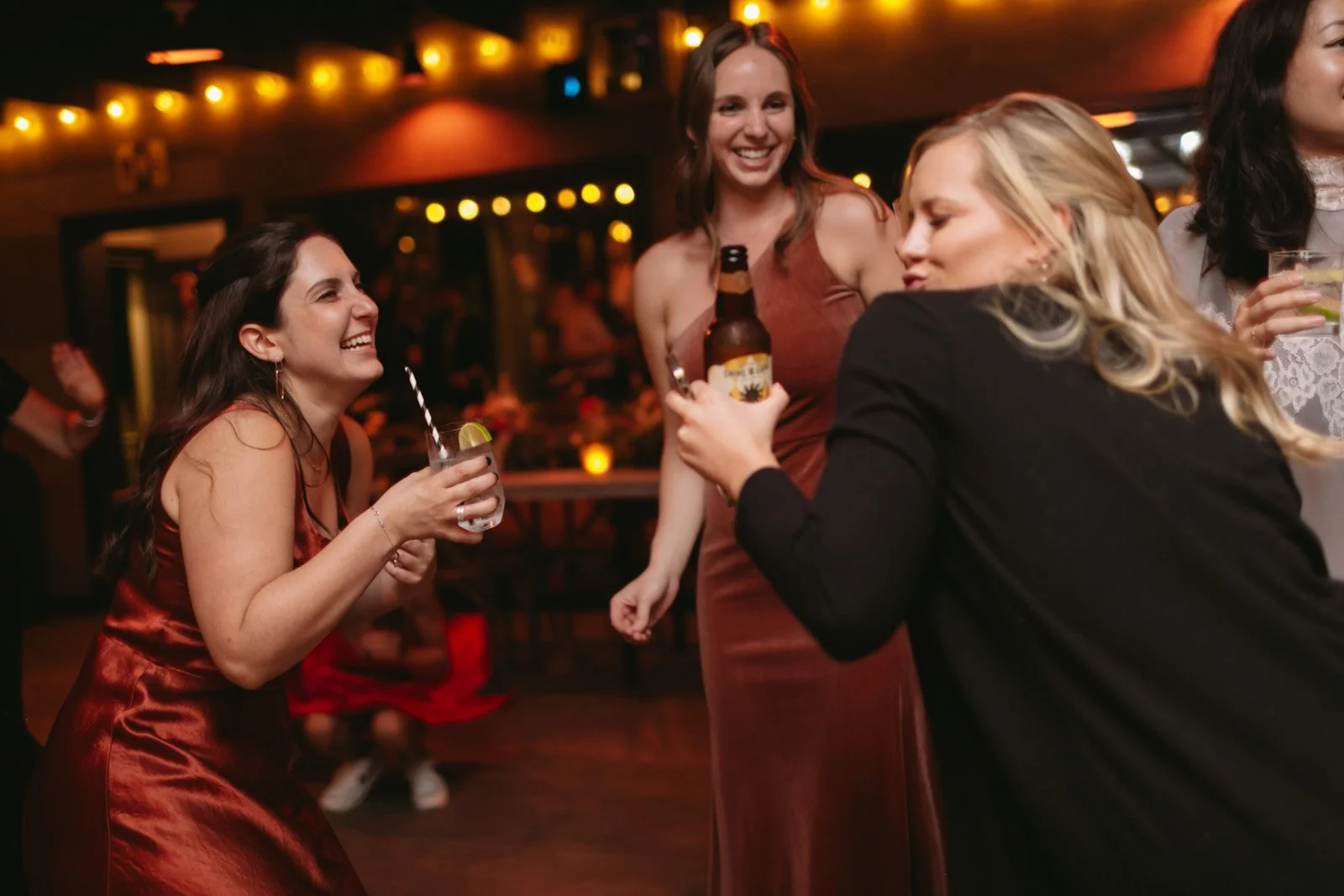 Four women enjoying drinks and conversation at a social gathering in a dimly lit indoor venue with warm lighting.