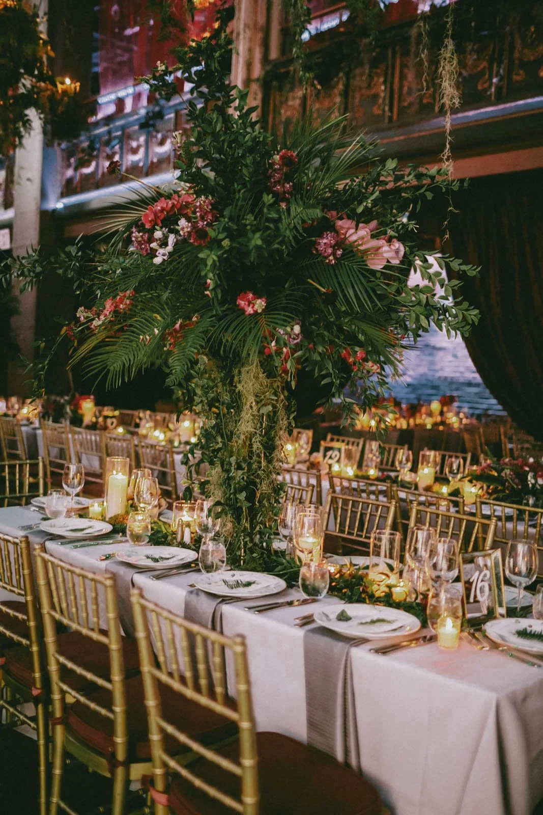 Elegant banquet table decorated with a tall floral centerpiece of greenery and pink flowers, surrounded by gold chairs, candles, wine glasses, and place settings in a warm, dimly lit indoor venue.