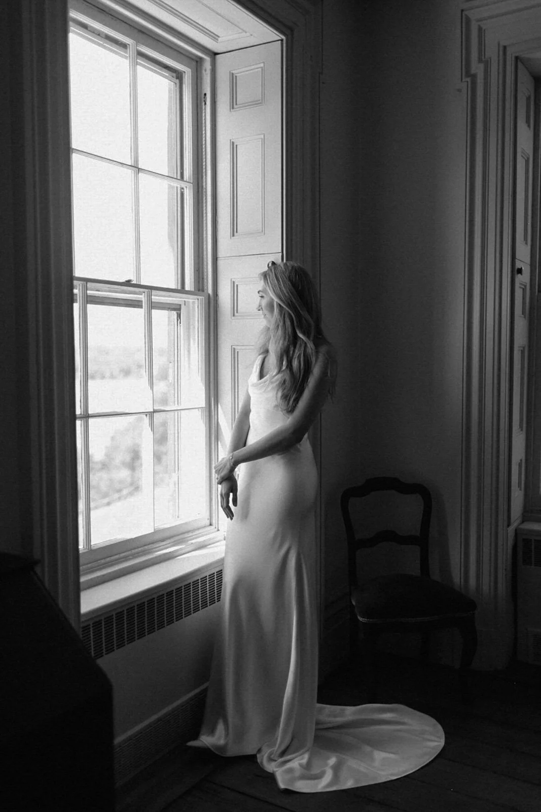 A woman in a wedding dress looking out a window in a room with wooden paneling and a chair.
