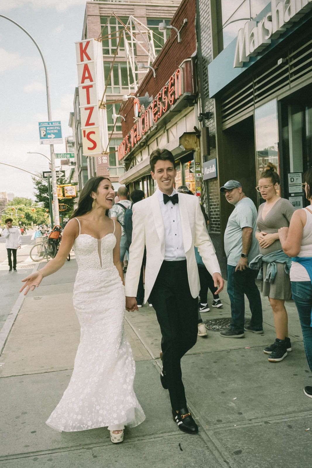 A couple dressed in wedding attire walking hand in hand on a city sidewalk, surrounded by onlookers and storefronts, including a pizza shop.
