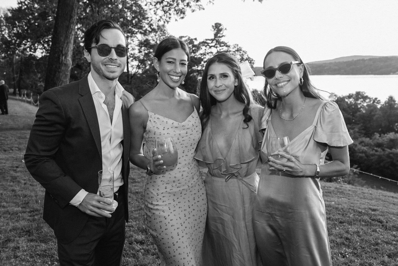 Four smiling people, two men and two women, standing outdoors near a body of water, holding drinks, dressed in semi-formal attire.