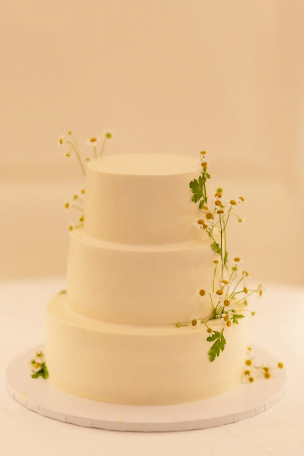 A three-tiered white cake decorated with small white and yellow flowers and green leaves.