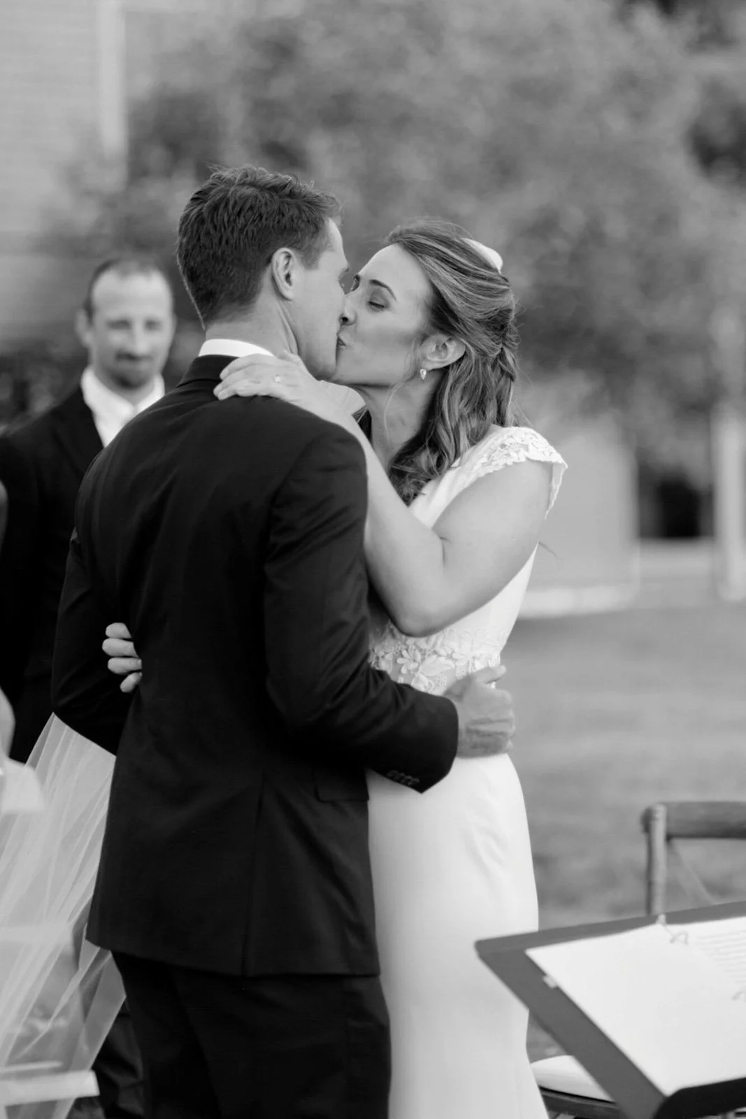A black and white photo of a bride and groom kissing during their wedding ceremony, with an officiant in the background.