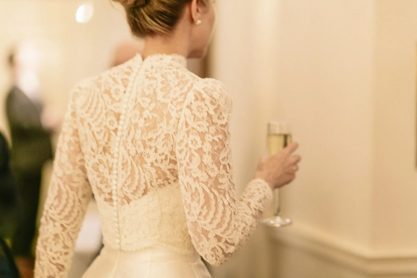 A woman in a cream lace dress holding a glass of champagne at an indoor social event.