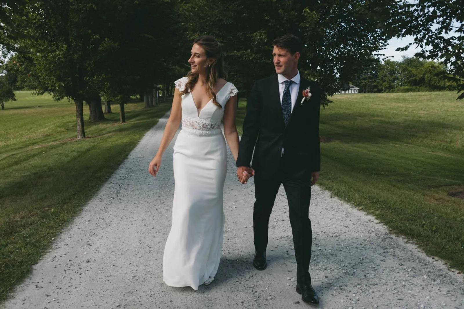 A bride and groom walking hand in hand on a gravel path surrounded by green grass and trees at Court Hill in The Berkshires.