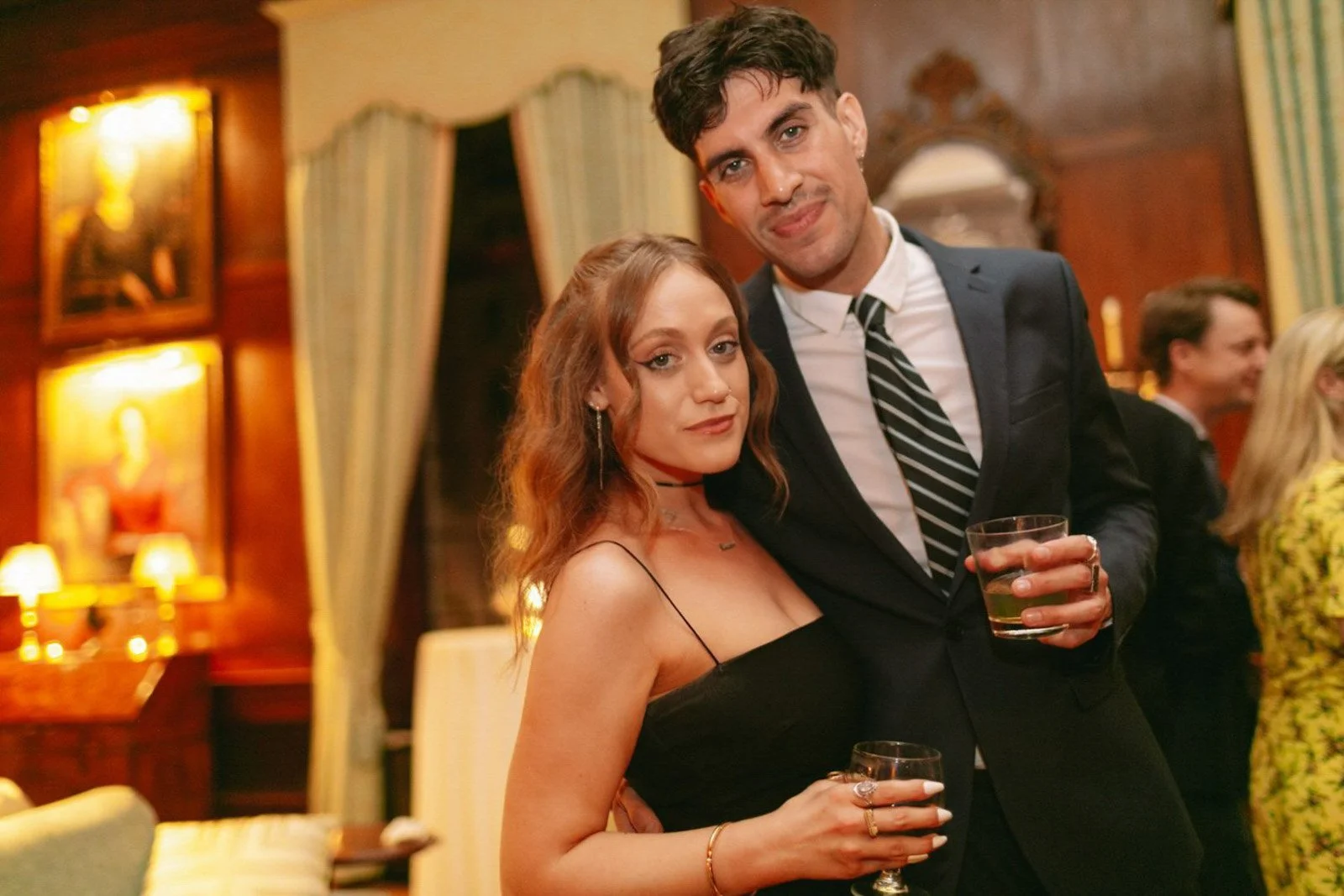 A young woman with wavy red hair and a young man with dark hair and a mustache at a social event, holding drinks and posing for a photo in an elegant, warmly lit room with vintage decor.