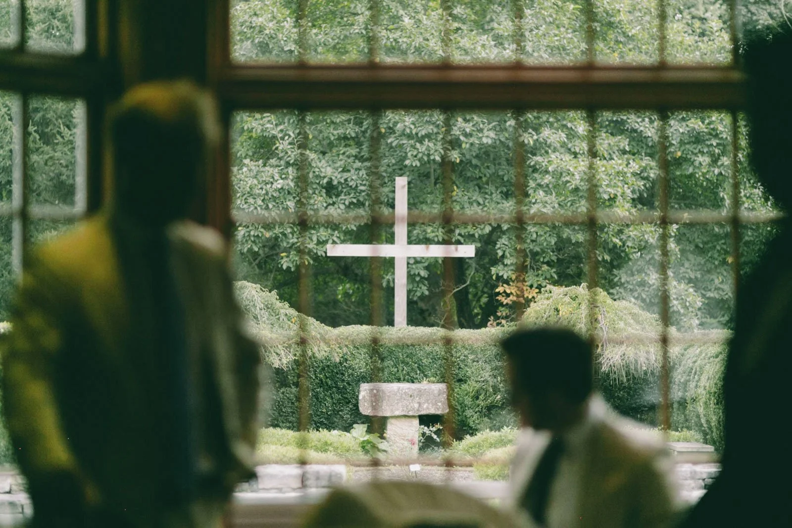 View through a window with metal bars, showing a cross monument and a bench outside surrounded by greenery.