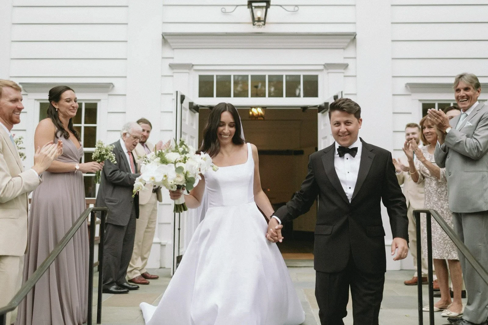 A newlywed couple holding hands and smiling as they walk out of Stanwich Church in Greenwich, CT, with family and friends applauding around them.